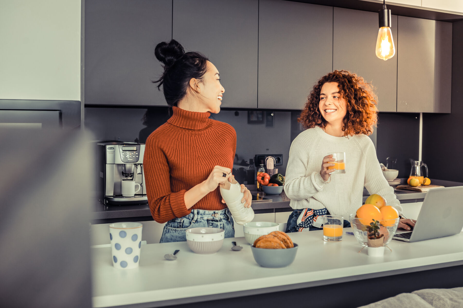 Two women laughing and enjoying food and beverages in a lively co-living community kitchen, reflecting a warm and communal atmosphere.
