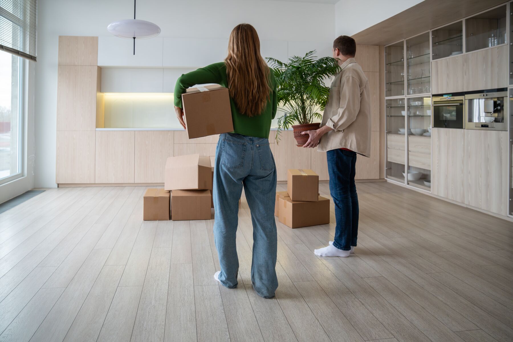 Rear view of new homeowners with boxes and houseplant in hands. Family couple taking things to another rented light apartment. Housewarming in mortgage apartment. Beginning of happy married life.