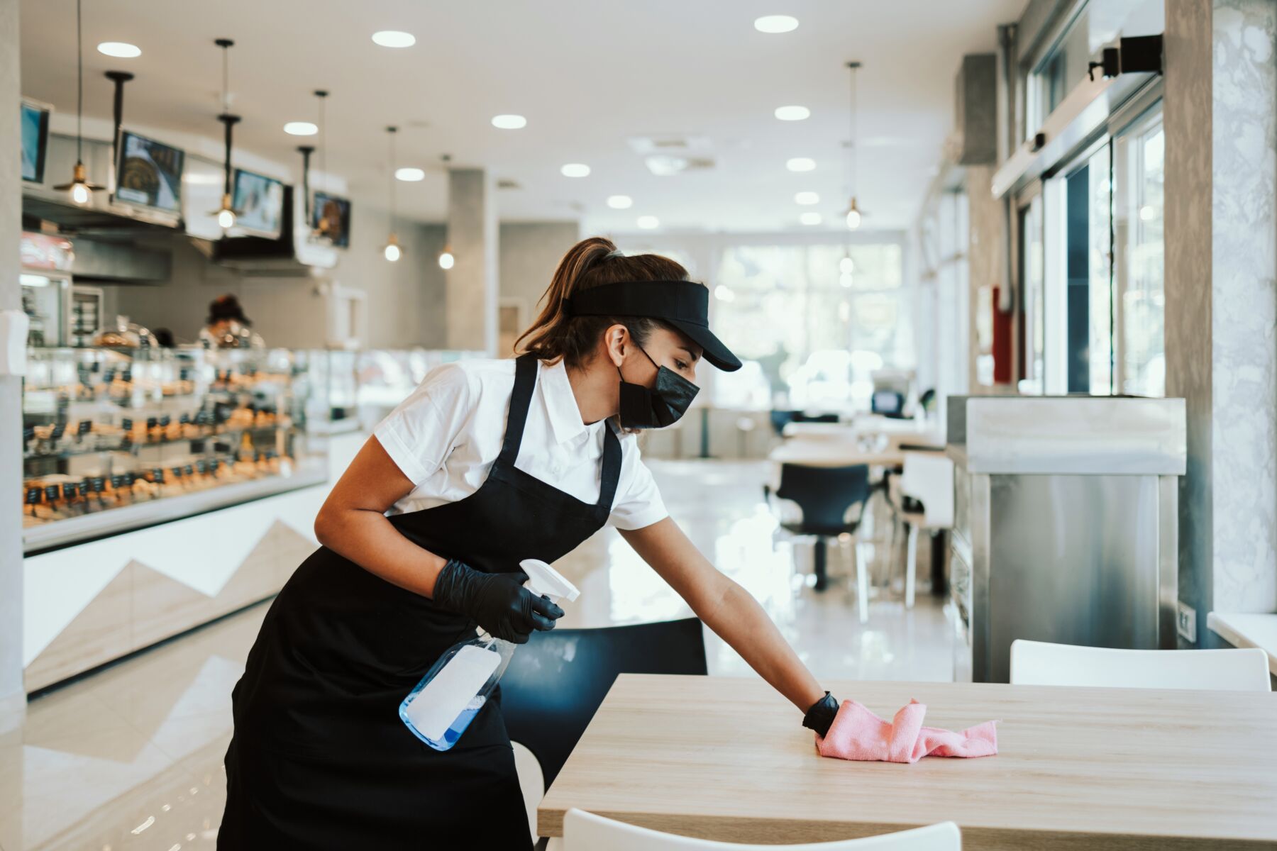 A woman with brown hair wears an apron and wipes down a table in a restaurant.