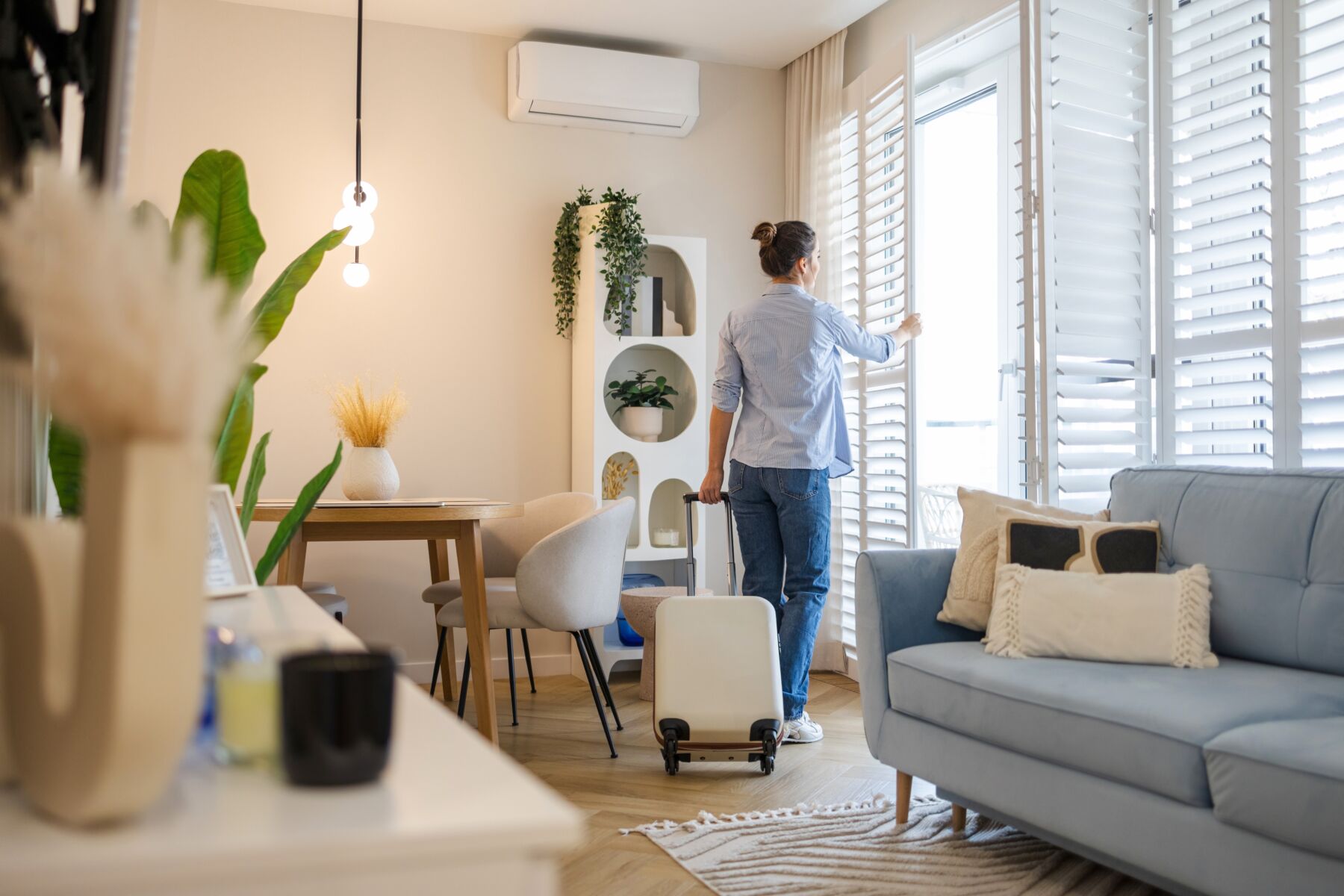 A woman walks around a dining room with a suitcase and looks out the window.