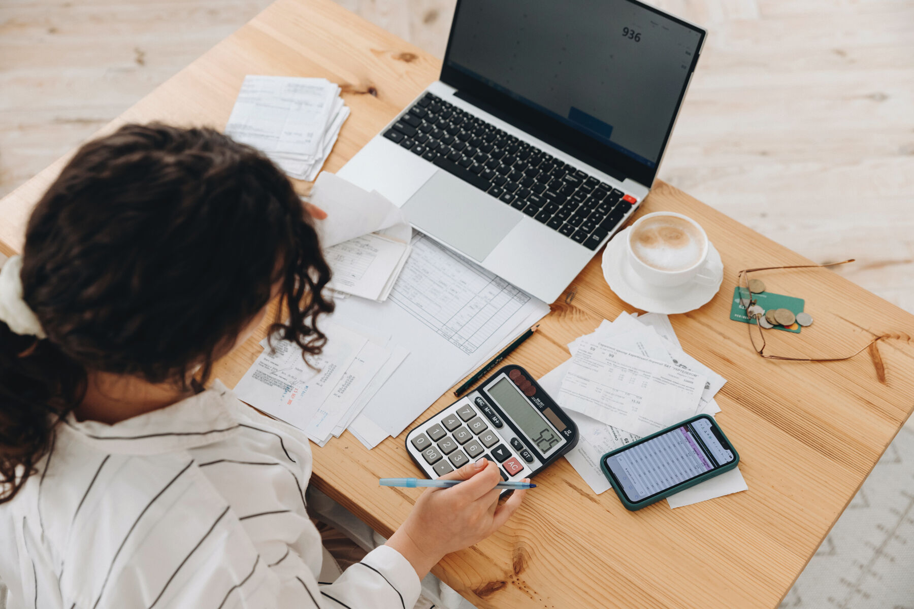Photo shows a woman working at home in the kitchen with financial papers, counting on a calculator, paying bills, planning a budget to save some money. Independent accounting, remote accountant.