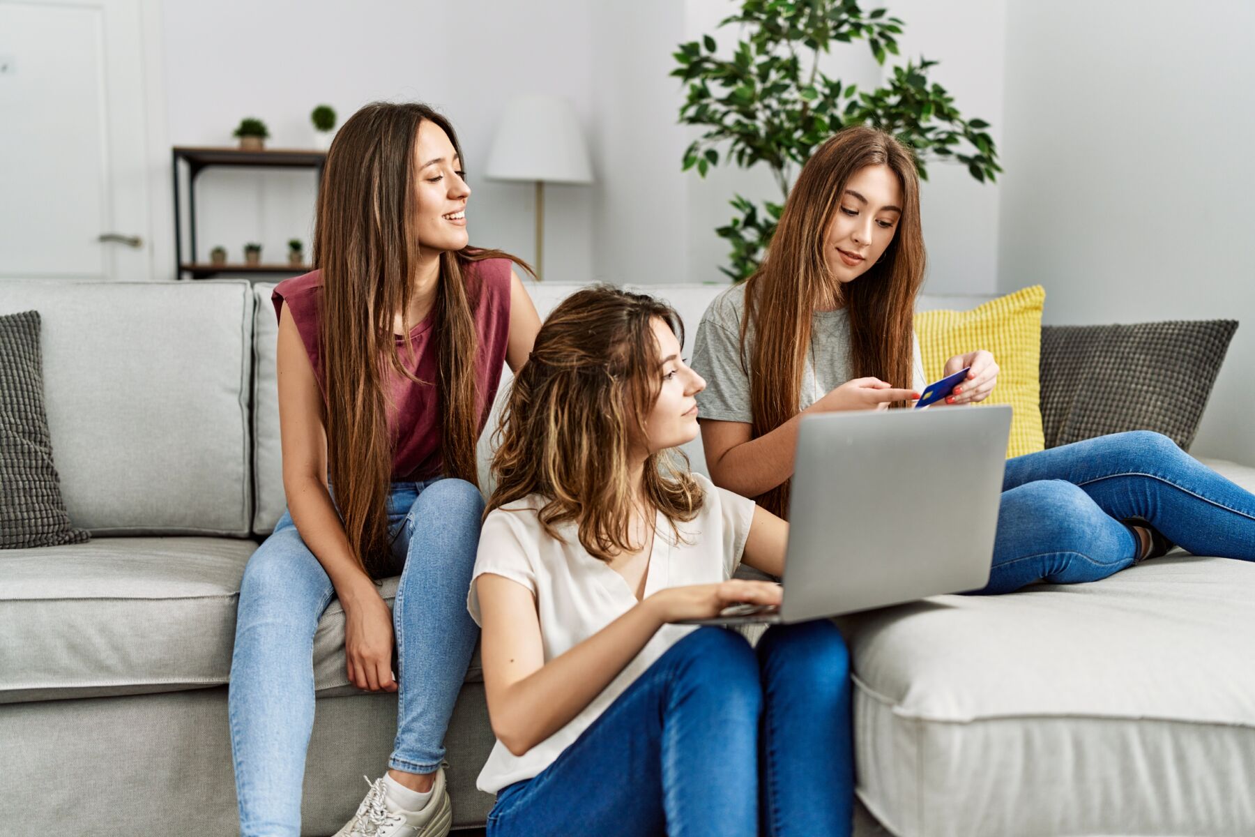 Photo shows three young PadSplit renters smiling happy using laptop and credit card sitting on the sofa at home.