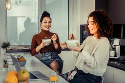 Two women hang out in a kitchen while eating food out of bowls.