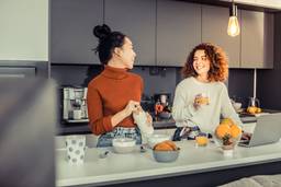 Two women laughing and enjoying food and beverages in a lively co-living community kitchen, reflecting a warm and communal atmosphere.