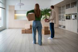 Rear view of new homeowners with boxes and houseplant in hands. Family couple taking things to another rented light apartment. Housewarming in mortgage apartment. Beginning of happy married life.