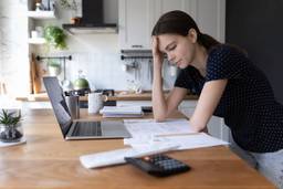 A Caucasian woman sits at a countertop and looks stressed while reviewing her bills.