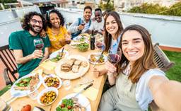 A group of friends eat a meal around a dining table and smile at the camera.