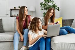 Photo shows three young PadSplit renters smiling happy using laptop and credit card sitting on the sofa at home.