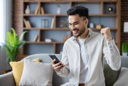 Photo shows man sitting on couch expressing excitement while looking at smartphone. Modern home interior with wooden shelves and plants in background. Concept of success, happiness, and achievement.
