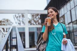 A confident female travel nurse in blue scrubs strides purposefully towards her new assignment. She carries a travel bag over one shoulder and holds a to-go coffee cup, symbolizing her readiness to take on a new challenge in a different location.