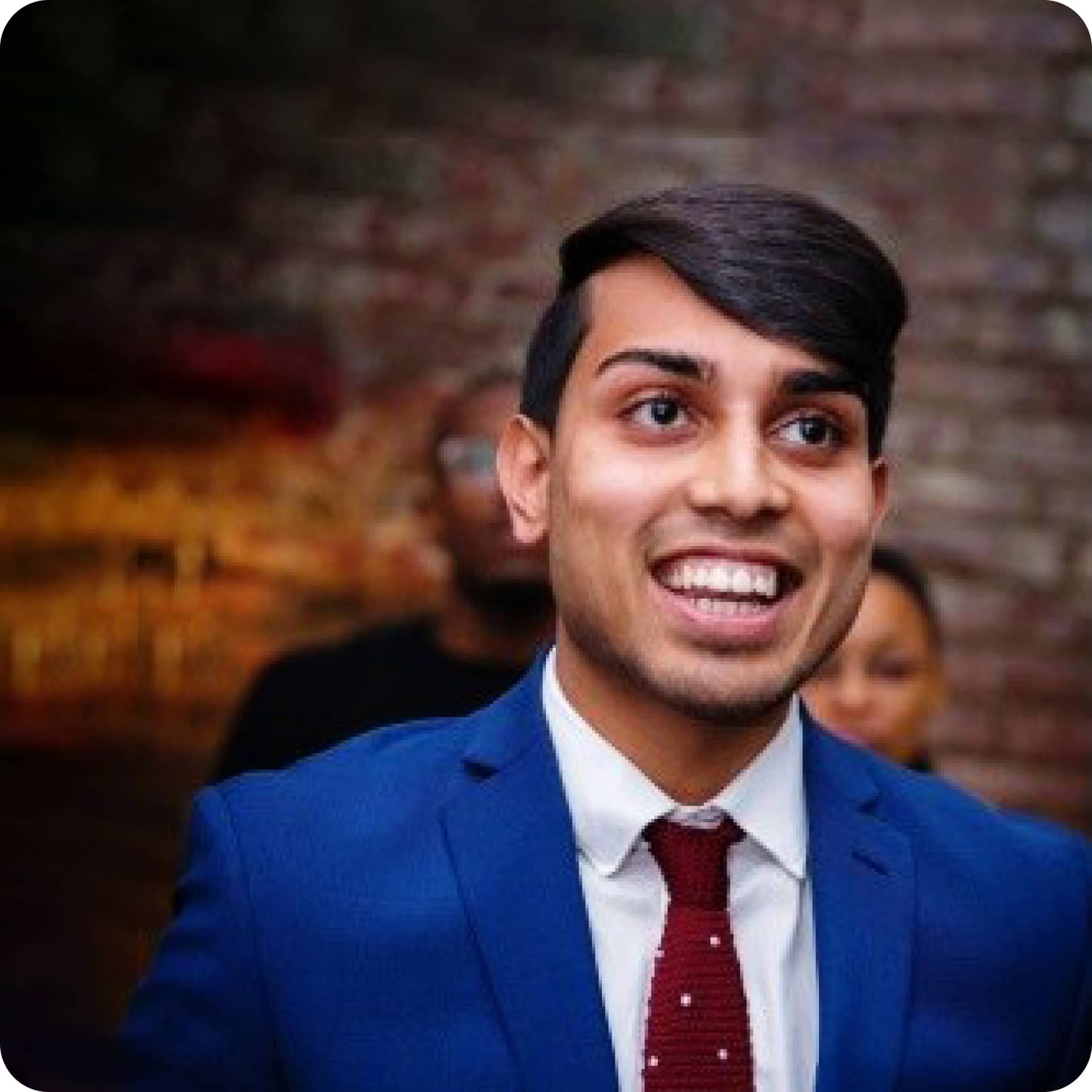 A young man with dark hair in a blue suit smiles in a crowded room.