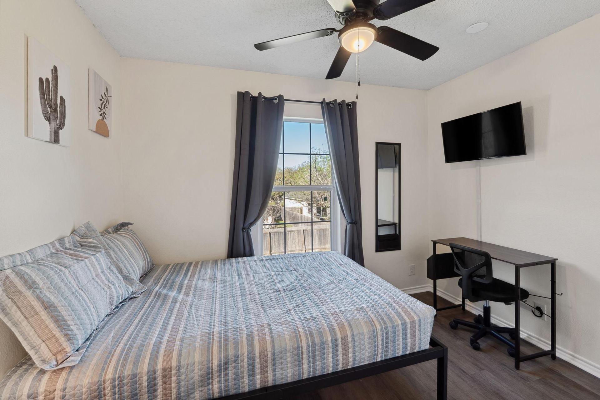 Bedroom with comfortable bed, a mini fridge, a TV, and a work desk. A mini closet & a ceiling fan.