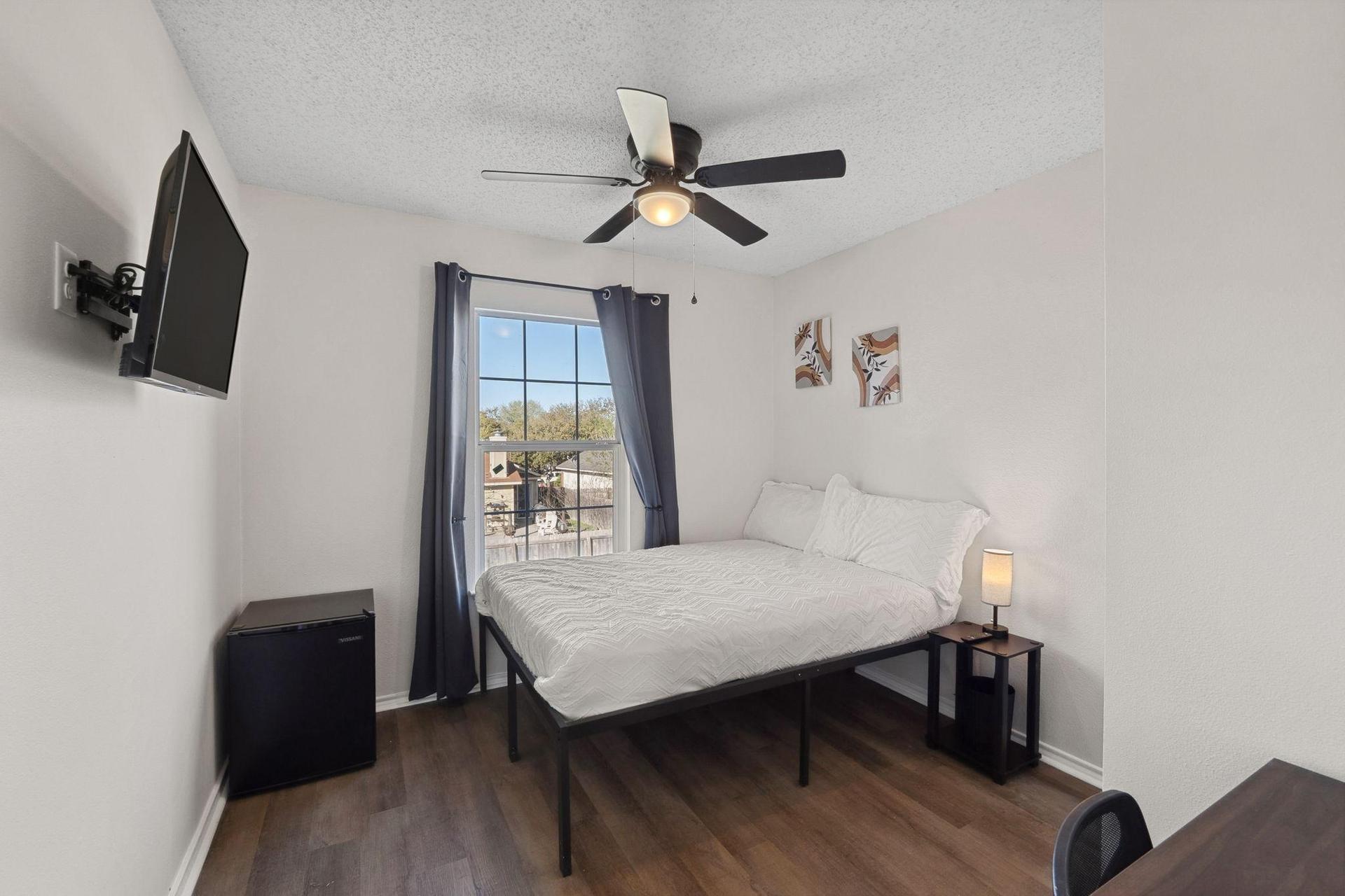 Bedroom with comfortable bed, a mini fridge, a TV, and a work desk. A mini closet & a ceiling fan.