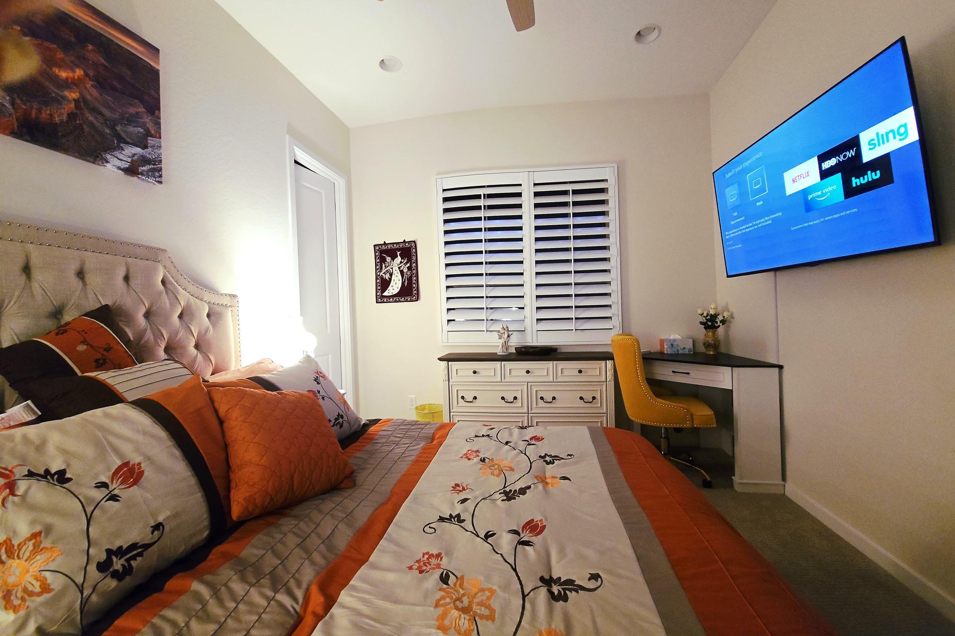 Cozy room with queen bed, tufted headboard, big smart TV, dresser, and a workspace desk with sandstone-colored chair.