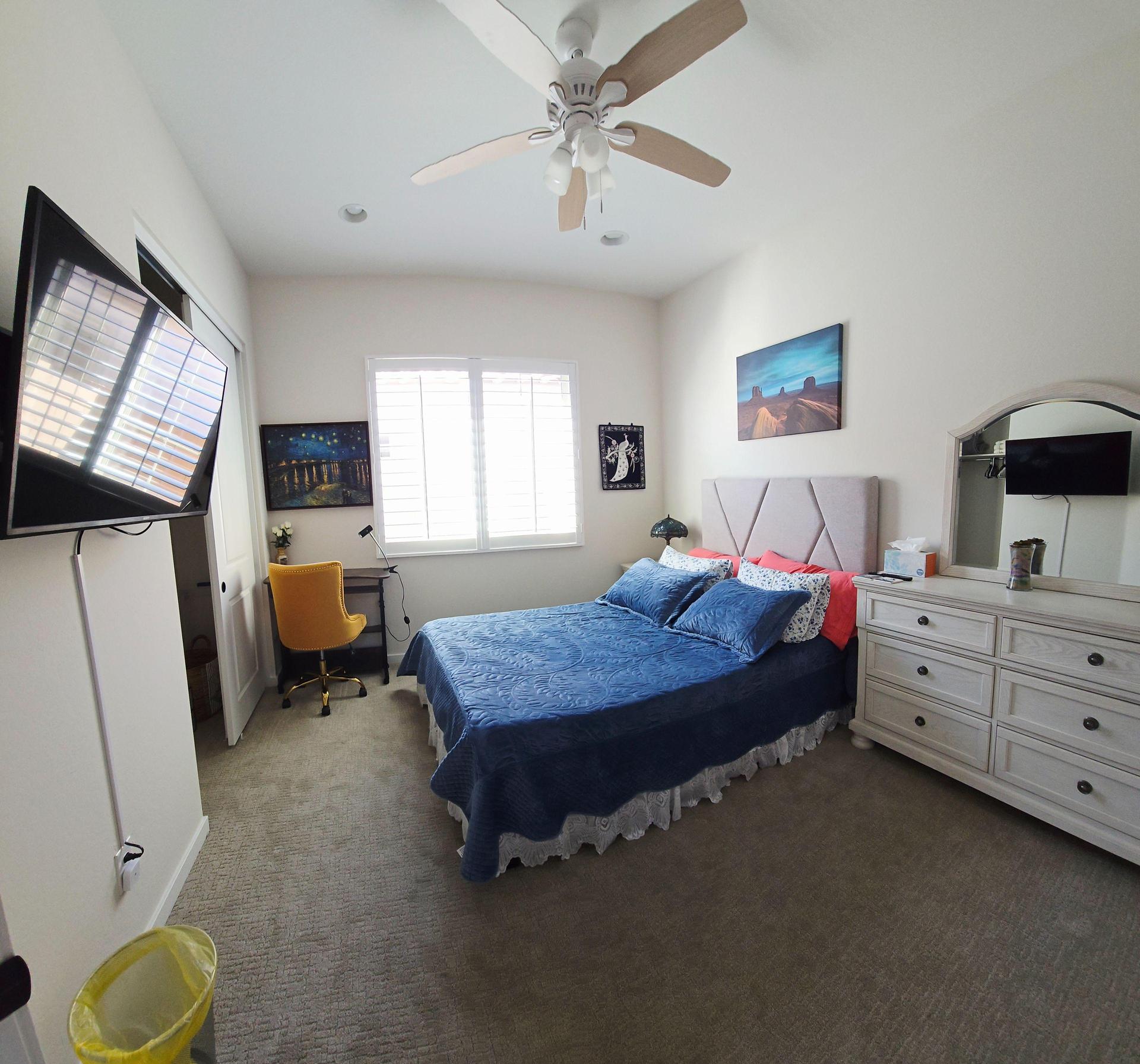 Queen bed with velvet blue cover, sand-colored pillows, desk with yellow chair, canyon art, TV, dresser, and shuttered window