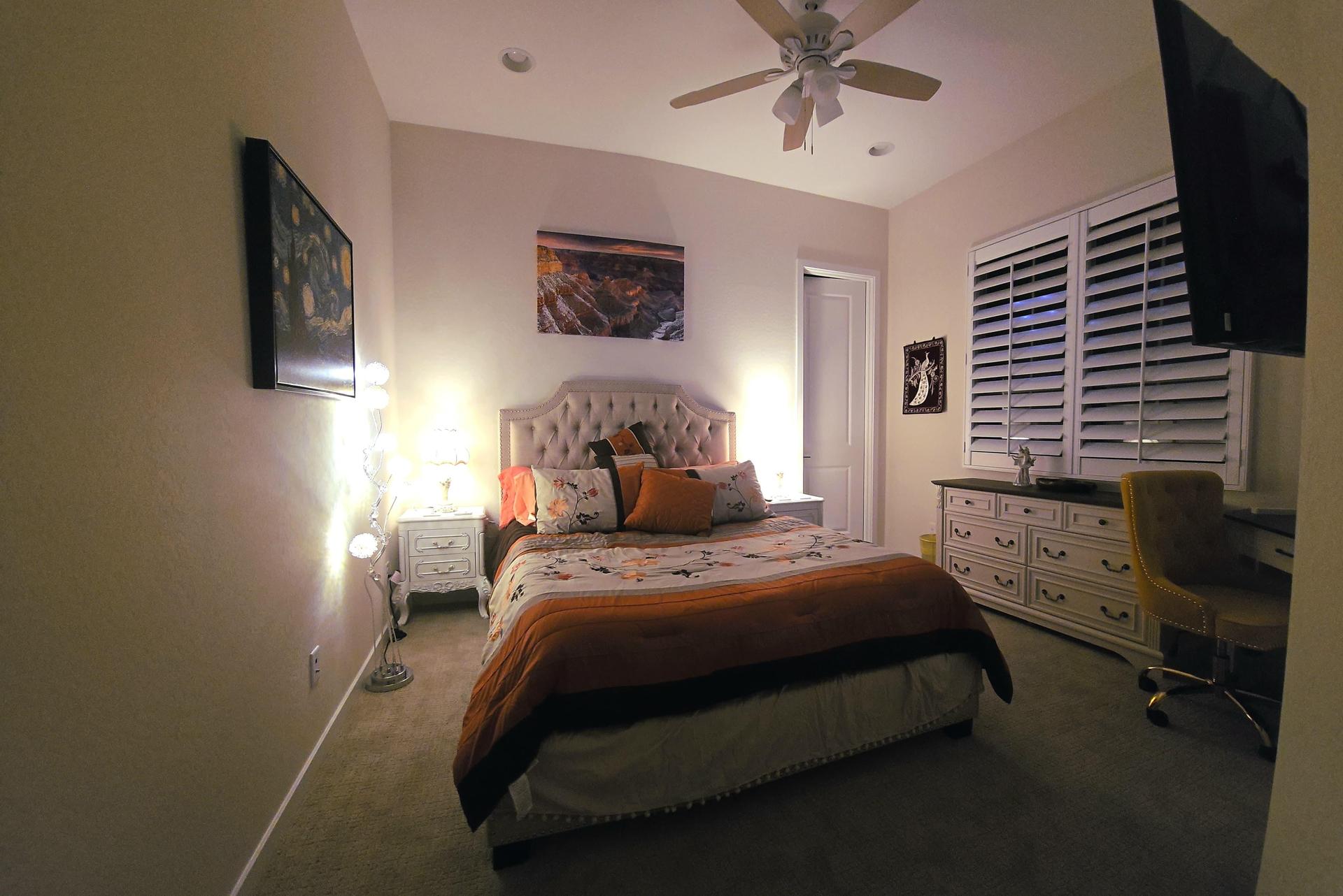 Cozy room with queen bed, tufted headboard, big smart TV, dresser, and a workspace desk with sandstone-colored chair.