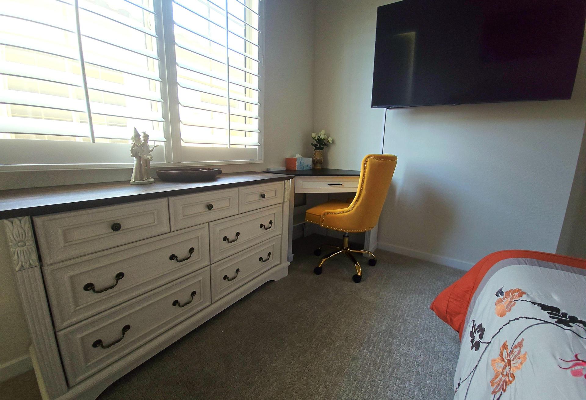 Cozy room with floral bedding, tufted headboard, big smart TV, dresser, and a workspace desk with sandstone-colored chair.